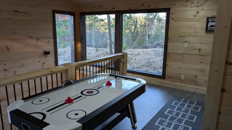 Air hockey table on the upstairs landing beside a stair rail and tall windows onto the woods