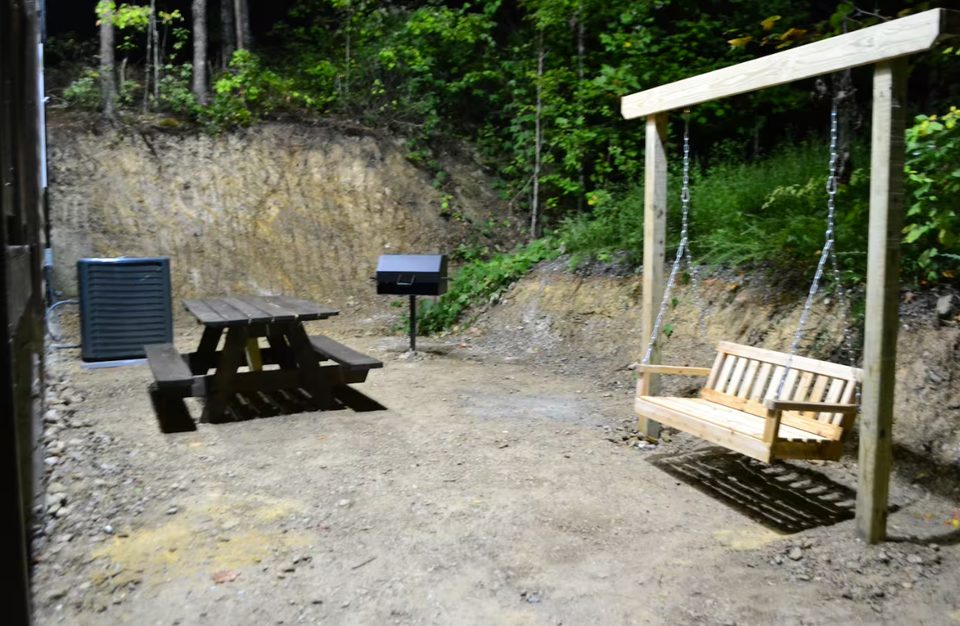 Charcoal grill on a post beside a picnic table, with a timber porch swing on the gravel below the cabin